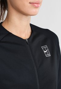 Mujer vestida con una chaqueta negra con cremallera y un logo deportivo blanco en el pecho, mostrada desde el cuello hasta la mitad del torso sobre un fondo liso.