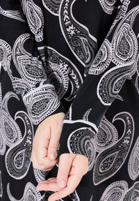 Mustaa kangasta, jossa on valkoinen ja harmaa paisley-kuvio. Pitkät hihat, joissa on kontrastivärit, näyttävät yksityiskohtaisia kuvioita ja sileää tekstuuria.