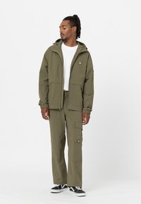 Giacca verde oliva con cappuccio e tasche frontali, abbinata a pantaloni cargo coordinati con tasche a bottone e con zip. Sneakers nere completano l'outfit.