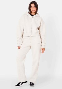 Ljusbeige huvtröja med dragkedja och ribbade muddarna, tillsammans med matchande träningsbyxor. Svarta sportskor med vita ränder. Mjuk, texturerad tyg.