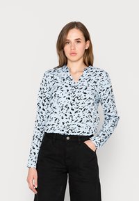 Hellbluse mit langen Ärmeln, die ein schwarzes abstraktes Muster, einen V-Ausschnitt und einen entspannten Schnitt aufweist. Kombiniert mit hoch taillierten schwarzen Hosen.