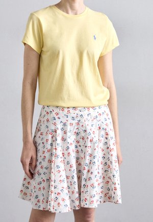 Kobieta ubrana w jasnożółtą koszulkę z krótkim rękawem z małym logo oraz białą spódnicę z czerwono-niebieskim kwiatowym wzorem i guzikami z przodu.