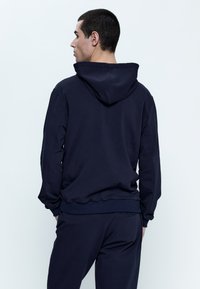 Felpa blu navy con cappuccio regolabile, polsini e orlo a coste. Realizzata in tessuto morbido, presenta una vestibilità rilassata e un dettaglio sulla cucitura posteriore.