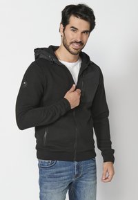 Felpa nera con zip, texture a costine, cappuccio regolabile con cordino e tasca sul petto con zip, indossata sopra una maglietta bianca e jeans blu.