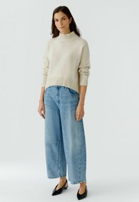Lichtbeige gebreide coltrui met geribbelde zoom, gecombineerd met lichte blauwe denim jeans met wijde pijpen, voorzien van voorzakken en versleten details.