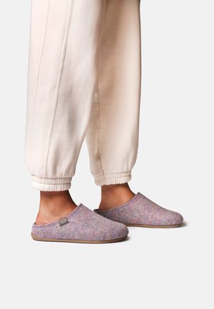 Personne portant des chaussons en feutre violet clair à enfiler et un pantalon de survêtement crème avec poignets, debout sur une surface blanche.