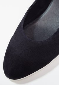 Gros plan d'une ballerine noire en daim avec bout arrondi et côtés échancrés sur fond blanc.
