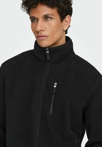 Muškarac s kovrčavom tamnom kosom, obučen u crnu jaknu od flisa s patentnim zatvaračem i visokom ovratnikom, s patentnim džepom na prsima, gleda u stranu.