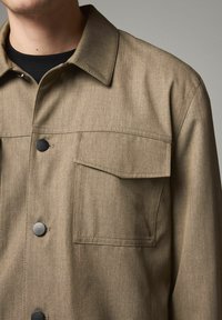 Jachetă button-up de culoare maro deschis, cu un finisaj texturat, două buzunare la piept și butoane din plastic negre. Dispune de un guler ascuțit și detalii cusute.