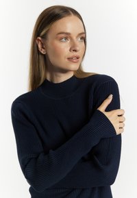 Granatowy sweter dzianinowy z prążkowanej tkaniny, o wysokim kołnierzu, długich rękawach i dopasowanych mankietach. Miękka faktura i luźny krój.
