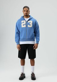 Blauwe zip-up hoodie met een witte versleten nummer "23," gedragen over een wit shirt. Zwarte korte broek, witte sokken en zwarte schoenen met roze veters.