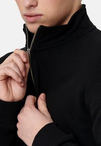 Czarna bluza z zamkiem błyskawicznym i wysokim kołnierzem, z ribbowe mankiety i widocznym srebrnym zamkiem, trzymana rękami w pobliżu dekoltu. Gładka tekstura.