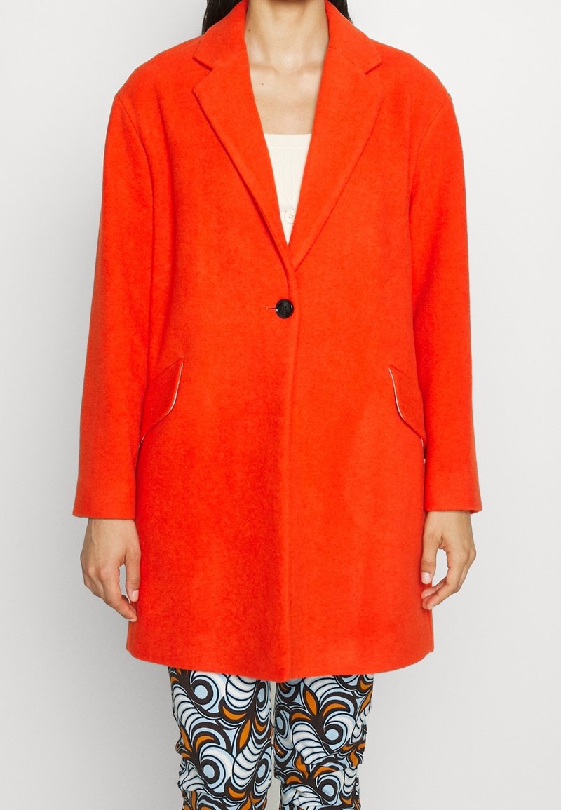 Donna che indossa un cappotto arancione brillante con un solo bottone sopra pantaloni neri, bianchi e arancioni a motivi, in piedi contro uno sfondo chiaro e uniforme.