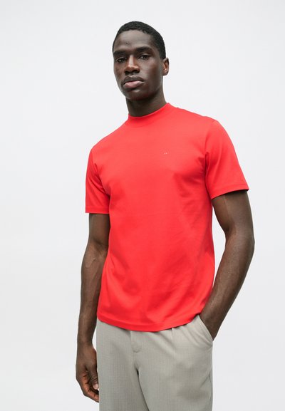 Νεαρός Μαύρος άνδρας φοράει ένα φωτεινό κόκκινο κοντομάνικο πουκάμισο και ανοιχτό γκρι παντελόνι, στέκεται με το ένα χέρι στην τσέπη του μπροστά σε μια απλή γκρι φόντο.