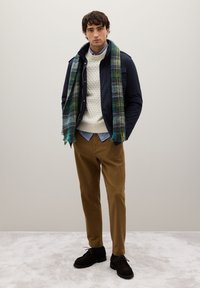 Giacca imbottita blu navy, maglione crema a trecce, camicia celeste con bottoni, pantaloni marroni e scarpe di camoscio nero. Sciarpa a quadri multicolore drappeggiata.