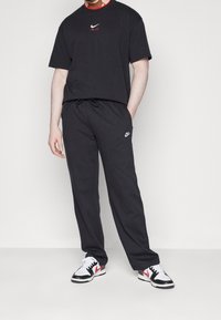 Uomo che indossa una maglietta nera Nike Air, pantaloni felpati neri Nike e sneakers Nike bianche, nere e rosse, in piedi contro uno sfondo bianco.