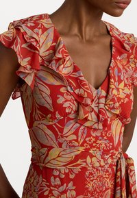 Robe florale rouge avec des manches courtes à volants, un décolleté en V profond et une ceinture à nouer à la taille. Le tissu a une finition texturée avec des accents pastel.
