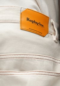 Μπεζ ύφασμα με πορτοκαλί δερμάτινη λεπτομέρεια που φέρει την ετικέτα "Replay Inc." και αντίθετες ραφές. Διαθέτει υφή στην επιφάνεια και λεπτομέρειες αντίκτυπου.