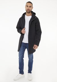 Chaqueta negra impermeable con capucha, cierre frontal de cremallera y bolsillos laterales. Llevada sobre una camisa blanca, combinada con jeans azules y zapatillas blancas.