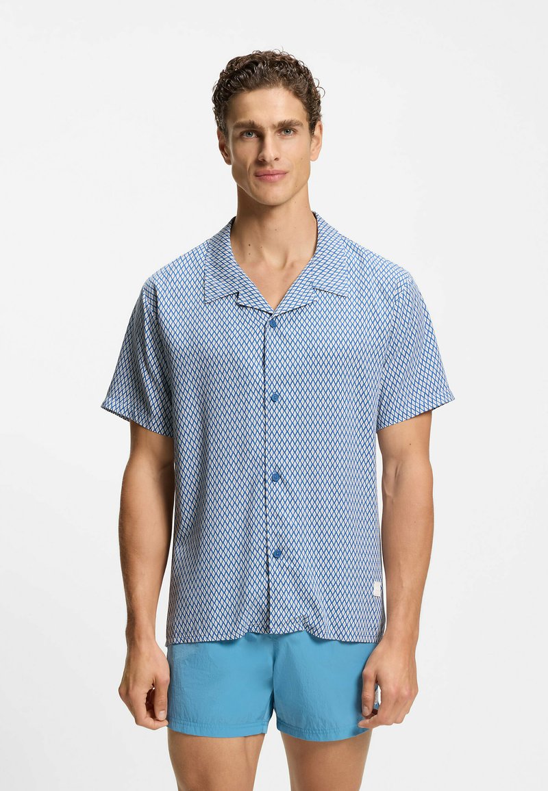 Uomo con capelli ricci che indossa una camicia a maniche corte blu e bianca con motivi a bottoni e pantaloncini azzurri uniformi, in piedi contro uno sfondo bianco.