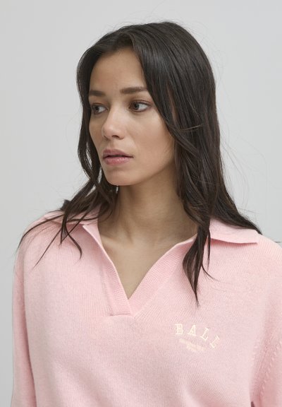 Svijetloružasti pulover s V-izrezom i ovratnikom. Ima teksturirani pleteni materijal i izvezeni detalj "BALL" na prsima.
