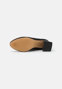 Zapato de tacón alto de ante negro con puntera afilada, suela de goma texturizada y tacón cuadrado. La suela es de color beige con estrías horizontales.