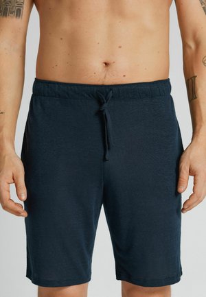 Person uden skjorte iført mørkeblå shorts med snøre, der viser overkrop, arme med tatoveringer, og hænder afslappet langs siderne mod en ensfarvet baggrund.