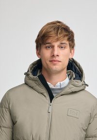 Giacca puffa beige con cappuccio, dotata di zip frontale, logo sul petto e tessuto testurizzato. Indossata sopra un maglione grigio chiaro e una camicia bianca con colletto.