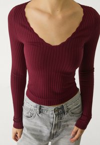 Haut bordeaux à manches longues côtelé avec un décolleté en V festonné, design ajusté et texture légère, associé à un jean gris clair.