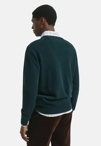 Dunkelgrüner Strickpullover mit einem gerippten Saum, taillierter Passform, getragen über einem hellen karierten Hemd. Weiche Textur, lange Ärmel und ein runder Ausschnitt.