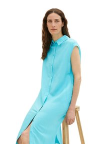 Ein langes, ärmelloses Knopfdress aus leuchtend türkisfarbenem Stoff mit glatter Textur und einem seitlichen Schlitz, ergänzt durch einen Holzstuhl.