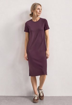 Frau mit blondem Haar in einem tiefen Dutt, trägt ein lila Midikleid mit kurzen Ärmeln und braune Sneaker, steht vor einer hellen Wand.