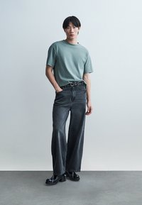 Mladý človek s krátkymi čiernymi vlasmi stojí, oblečený v tyrkysovom tričku, širokých vyblednutých čiernych džínsach, čiernom opasku a čiernych platformových topánkach.