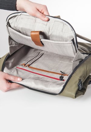 Mains tenant un sac vert olive ouvert montrant un intérieur beige doublé avec des poches zippées et des poches plates ainsi qu’un détail de sangle en cuir.