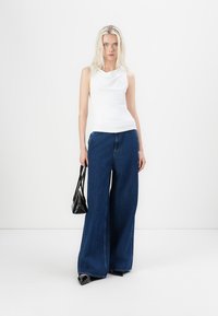 White sleeveless top with cowl neckline, paired with dark blue wide-leg denim pants. Model holds a small black handbag. Neutral background.