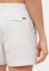 Costume da bagno di un beige chiaro, realizzato in materiale liscio e leggero. Caratterizzato da una vita elastica, tasche laterali e una tasca posteriore con etichetta del brand.