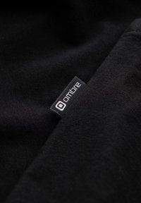 Schwarzer Stoff, der eine Nahaufnahme eines Etiketts eines Kleidungsstücks zeigt. Das Etikett trägt den weißen Text "ombre" mit einem minimalistischen Logo darüber.