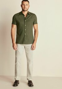 Camicia a maniche corte di colore verde oliva con tasca, design a bottone, abbinata a pantaloni beige chiaro e scarpe marroni scure, in piedi su uno sfondo neutro.
