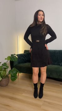 Svart långärmad klänning med markerad midja och ärmar som når axlarna, utrustad med ett metalliskt spänne på bältet. Knälång fåll, kombinerad med svarta ankelstövlar.