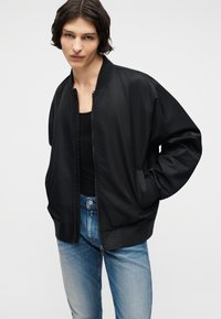 Νεαρό άτομο με σκούρα μαλλιά μέχρι τους ώμους, φορώντας μαύρο μπουφάν τύπου μπομπέρ πάνω από μαύρο τοπ και ανοιχτόχρωμο μπλε τζιν, με τα χέρια στις τσέπες του μπουφάν.