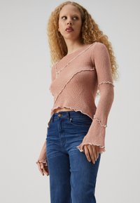 Svetlo roza pletena zgornja majica z razširjenimi rokavi in robovi z volančki, kombinirana s temno modrimi oprijetimi jeans hlačami.