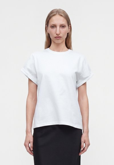 Woman with straight blond hair wearing a plain white short-sleeve shirt and a black skirt, standing against a white background.