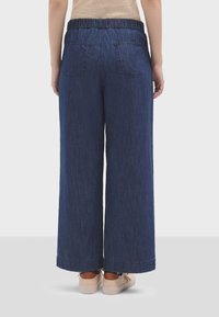 Pantaloni in denim blu scuro a gamba larga con vita elastica, due tasche posteriori e orlo pulito. Abbinati a scarpe chiare.