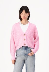 Rosa strikket cardigan med V-hals, tre store knapper og vide ermer, kombinert med lyseblå jeans med stjernesøm.