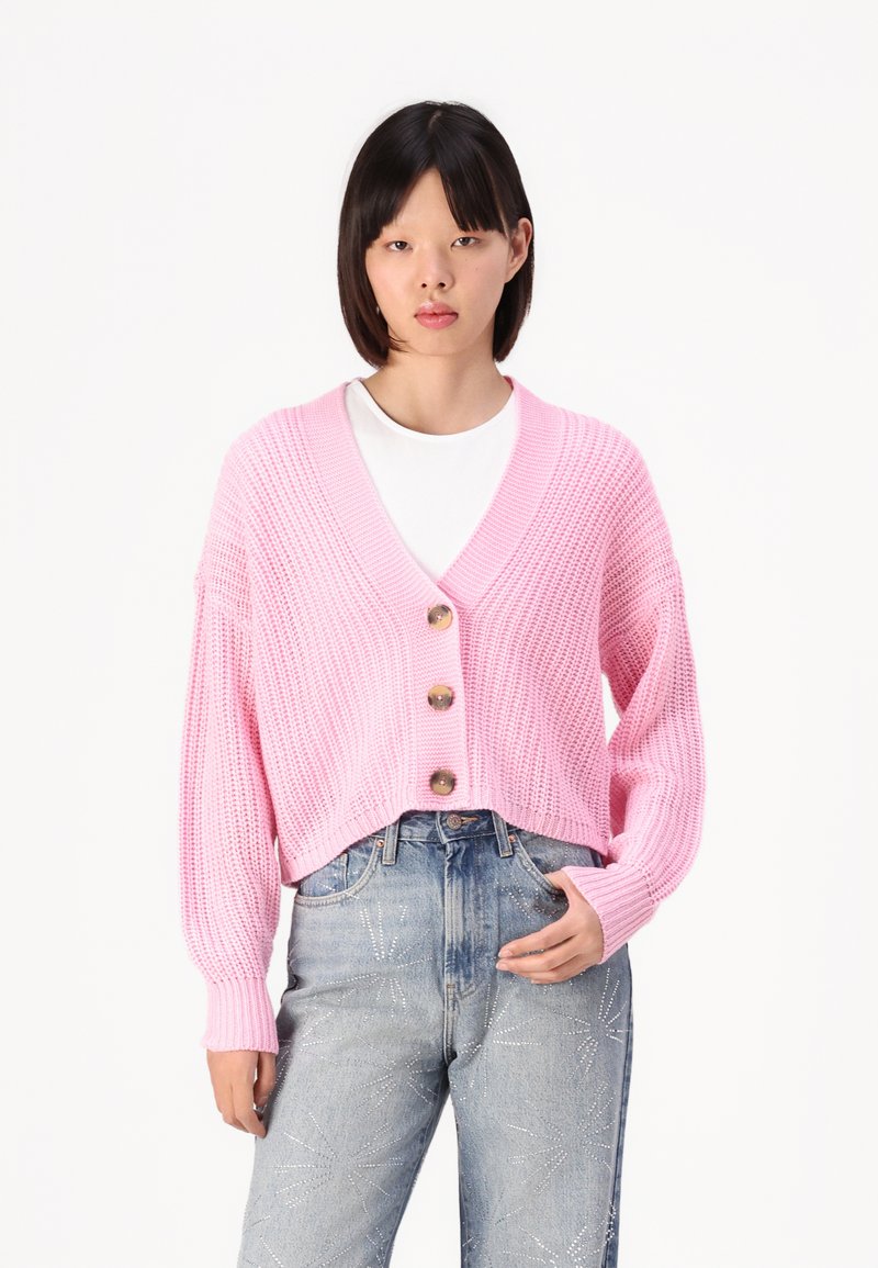 Rosa strikket cardigan med V-hals, tre store knapper og vide ermer, kombinert med lyseblå jeans med stjernesøm.