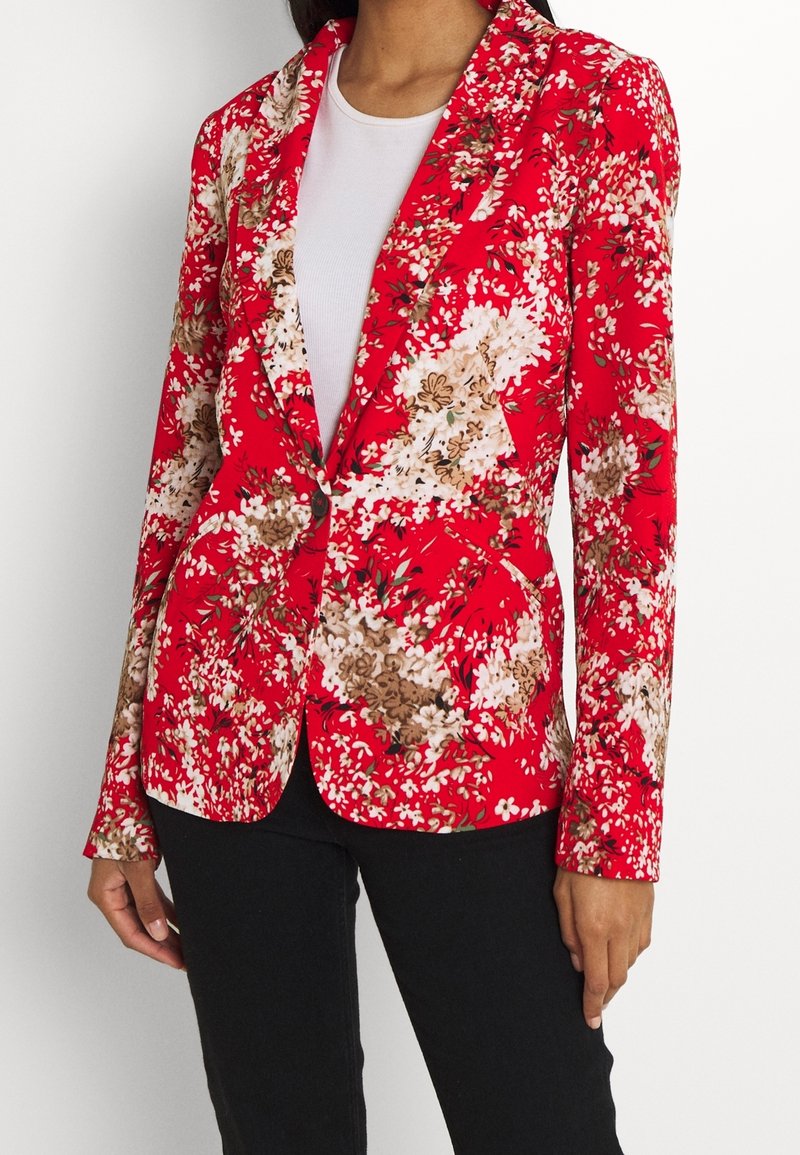 Femme portant un blazer rouge à fleurs blanches et beiges sur un top blanc et un pantalon noir, debout devant un fond uni.