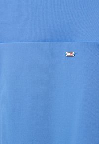 Top sportiv de culoare albastru deschis, realizat dintr-un material texturat. Are un mic logo al brandului cu accente roșii și albe pe partea stângă.