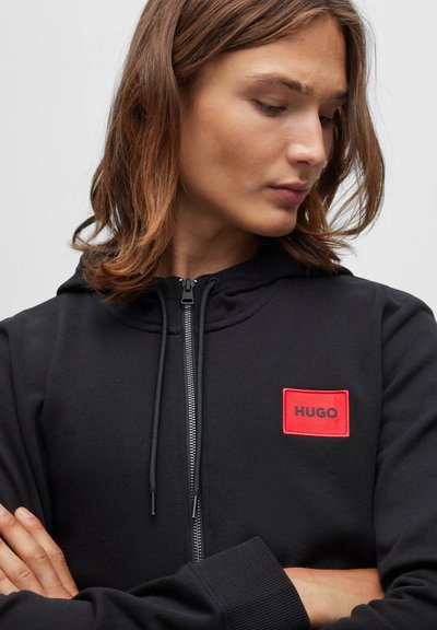 Jeune personne aux cheveux bruns mi-longs portant un sweat à capuche noir zippé avec un écusson du logo Hugo rouge sur la poitrine.