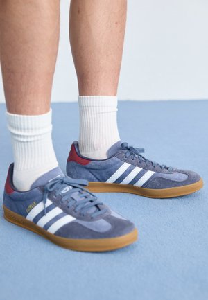 Personne portant des chaussettes blanches et des baskets en daim bleu avec des bandes blanches et des semelles en gomme, debout sur un sol bleu.