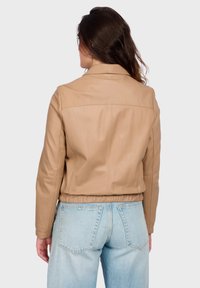 Frau mit langen dunklen Haaren, die eine beige Lederjacke und hellblaue Jeans trägt, steht mit dem Rücken zum Betrachter vor einem schlichten Hintergrund.